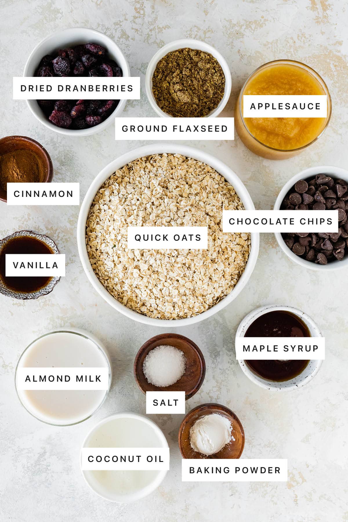 Ingredients measured out to make Oatmeal Breakfast Bars: dried cranberries, ground flaxseed, applesauce, cinnamon, quick oats, chocolate chips, vanilla, maple syrup, salt, almond milk, coconut oil and baking powder.