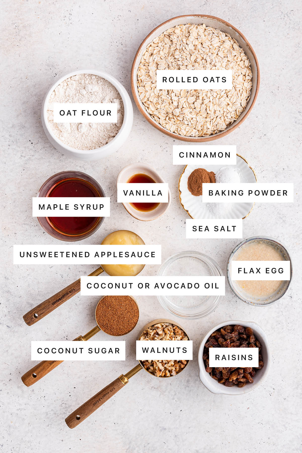 Ingredients measured out to make Oatmeal Breakfast Cookies: oat flour, rolled oats, maple syrup, vanilla, cinnamon, baking powder, sea salt, applesauce, coconut oil, flax egg, coconut sugar, walnuts and raisins.