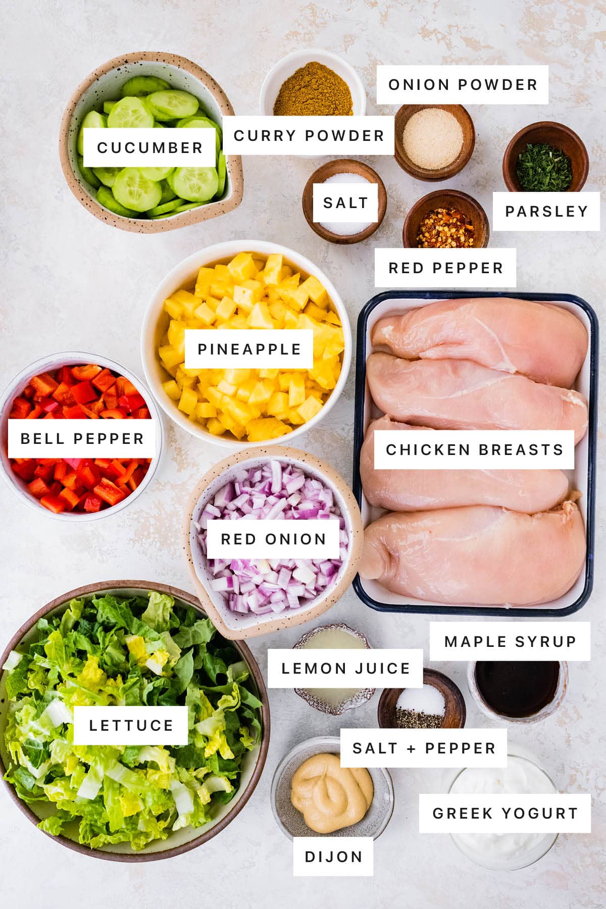 Ingredients measured out to make Thai Curry Chicken Salad: cucumber, curry powder, onion powder, salt, red pepper, parsley, pineapple, bell pepper, red onion, chicken breasts, lemon juice, maple syrup, lettuce, pepper, dijon and Greek yogurt.