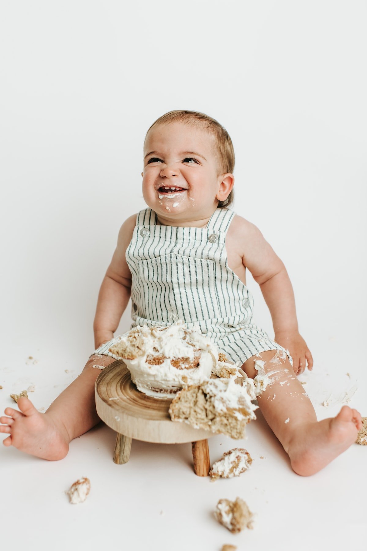 One year old baby boy in overalls smiling with smashed cake in front of him.