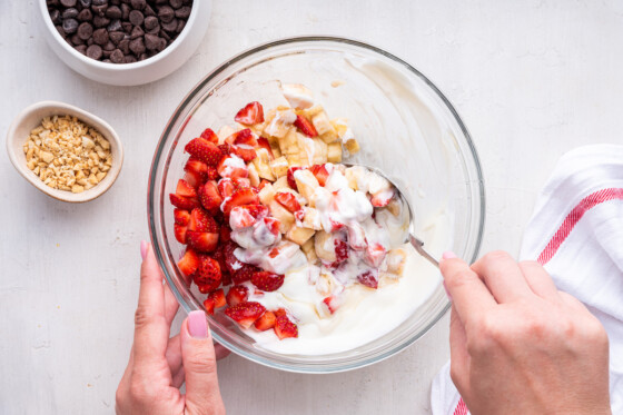 A woman's hand using a spoon to mix bananas, strawberries, and yogurt in a large glass bowl.