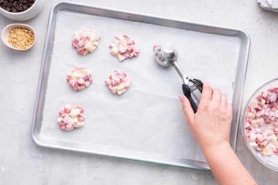 A woman's hand using a cookie scoop to form the yogurt clusters on a baking tray.