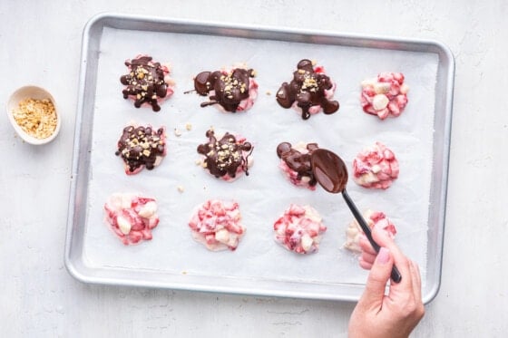 A woman's hand with a spoon, drizzling melted chocolate onto the strawberry banana yogurt clusters.