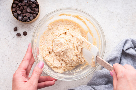 A woman's hand holding a bowl and using her other hand to fold the cottage cheese cookie dough with a silicone spatula.