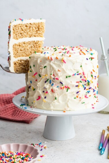 A slice of cake is removed from the full vanilla cake that sits on a white pedestal. Cake is decorated with vanilla frosting and rainbow sprinkles.