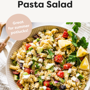 Greek pasta salad in a large white bowl with wooden serving spoons to the side.