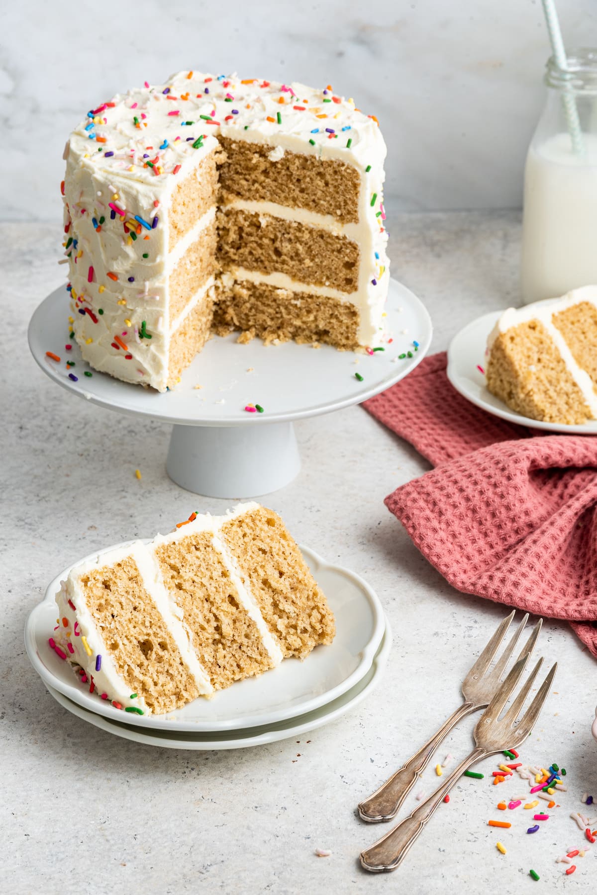 A full vanilla cake sitting on a white cake pedestal with slices removed. Slices are served in front of the cake on white plates.