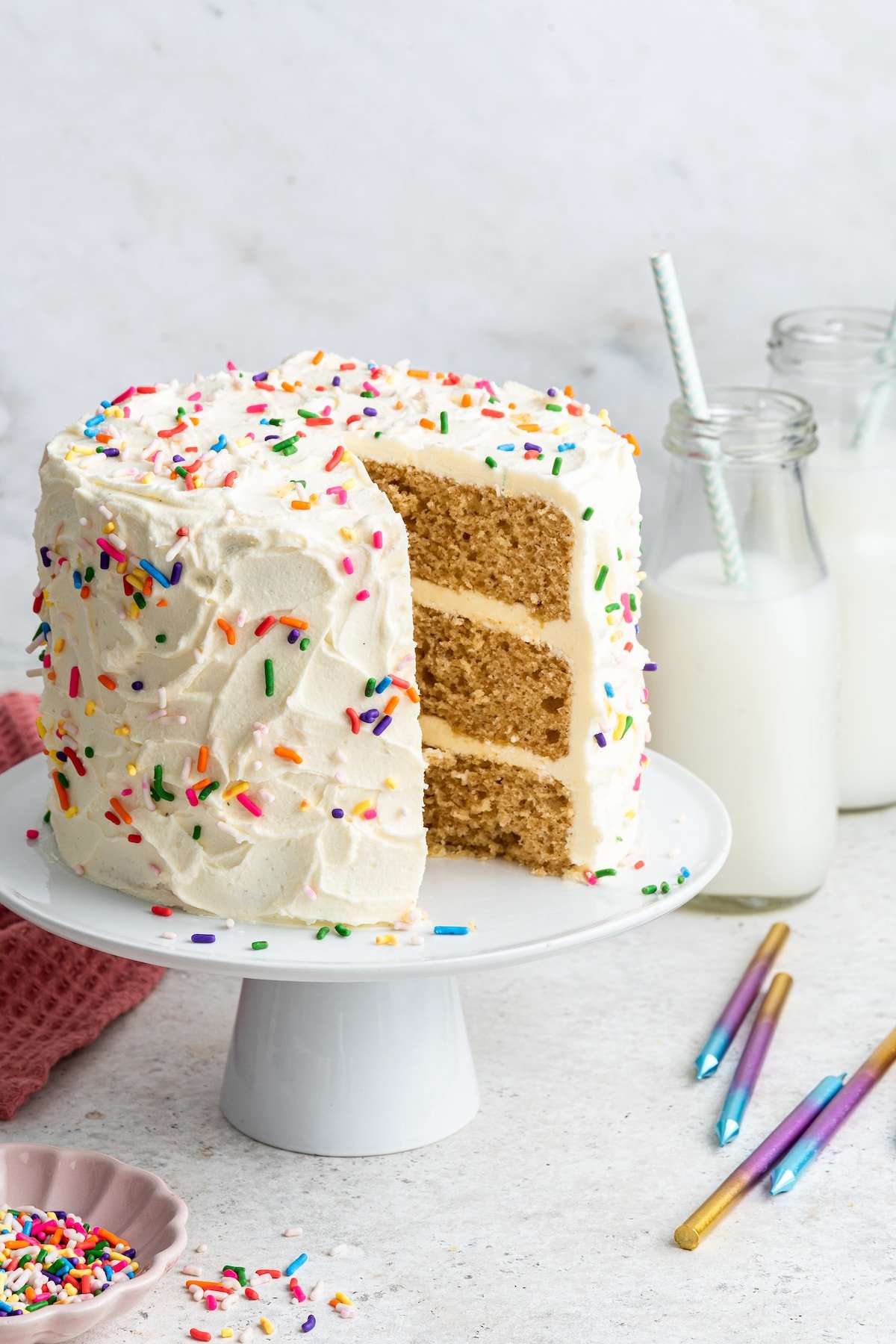 A vanilla cake frosted with rainbow sprinkles sitting on a cake pedestal with one slice of cake removed.