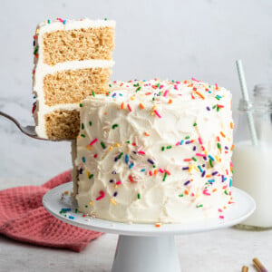A slice of cake is removed from the full vanilla cake that sits on a white pedestal. Cake is decorated with vanilla frosting and rainbow sprinkles.
