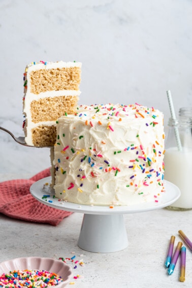 A slice of cake is removed from the full vanilla cake that sits on a white pedestal. Cake is decorated with vanilla frosting and rainbow sprinkles.