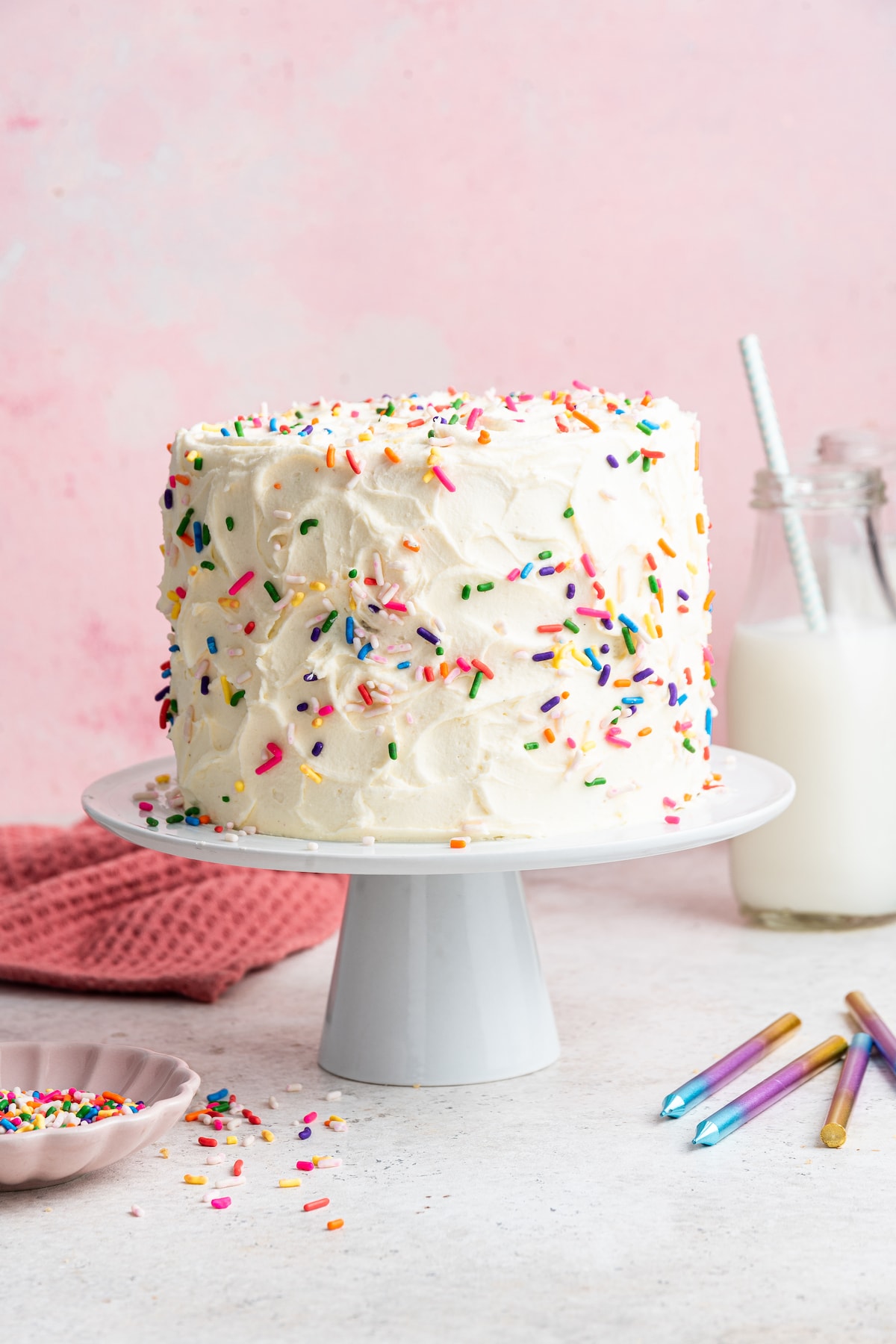 A vanilla cake frosted with rainbow sprinkles sitting on a cake pedestal.