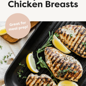 Three grilled chicken breasts on a grill pan with lemon slices and herbs.
