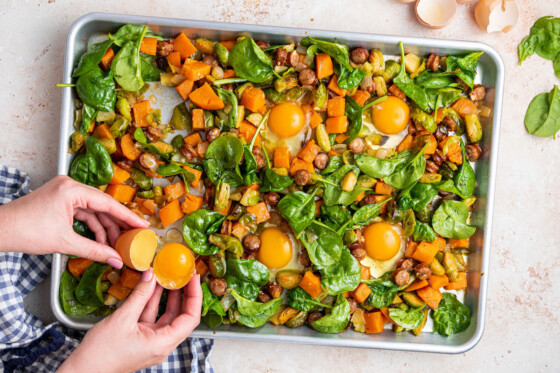 A woman's hand cracking an egg to place in a space on the baking tray full of the sweet potato hash.