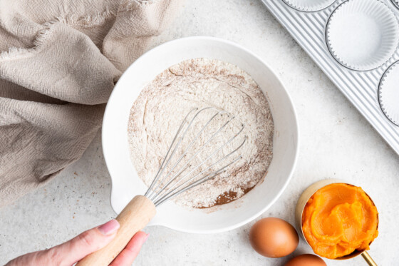 A woman's hand holds a metal whisk in a mixing bowl of flour, spices, baking powder, and baking soda.