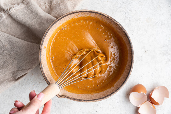 A woman's hand holds a metal whisk, using it in a large mixing bowl of wet ingredients for the sweet potato muffins.