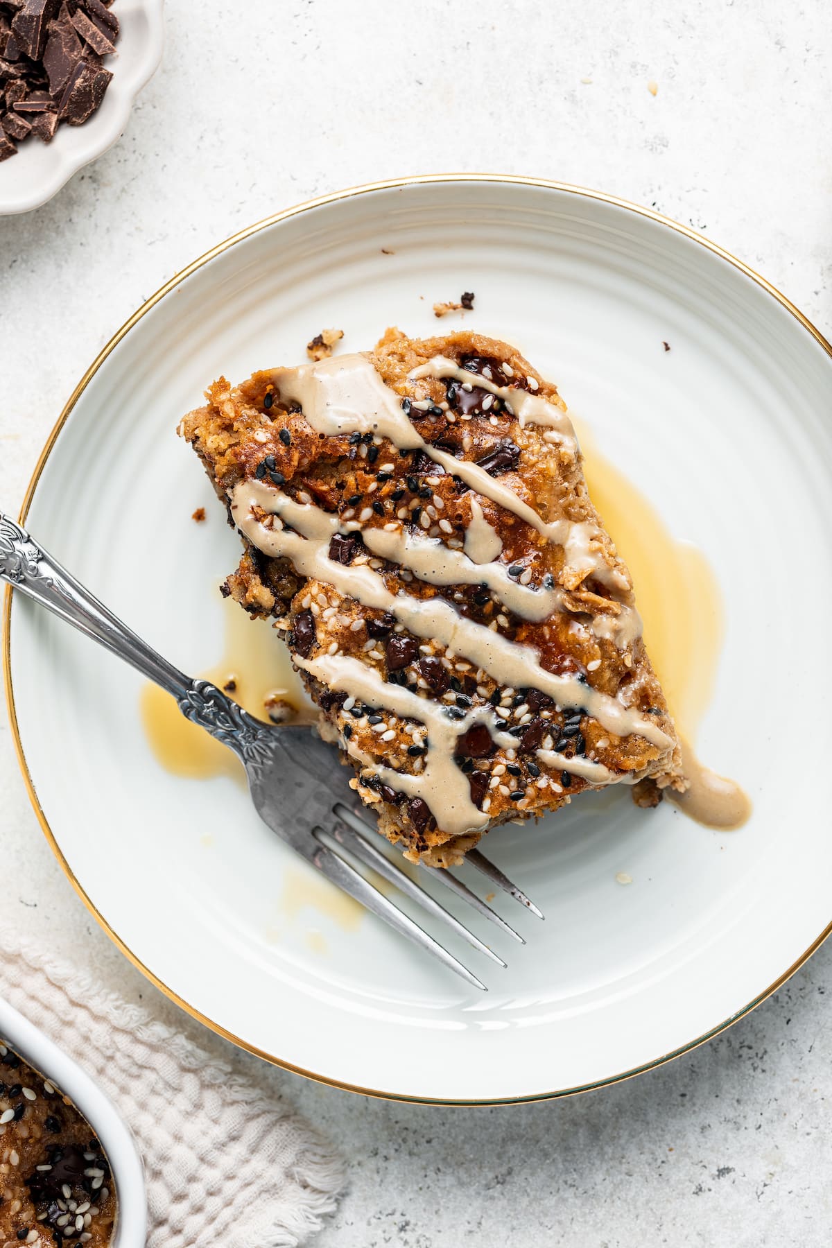 A piece of tahini baked oatmeal on a white plate with a metal fork, topped with a drizzle of tahini and maple syrup.