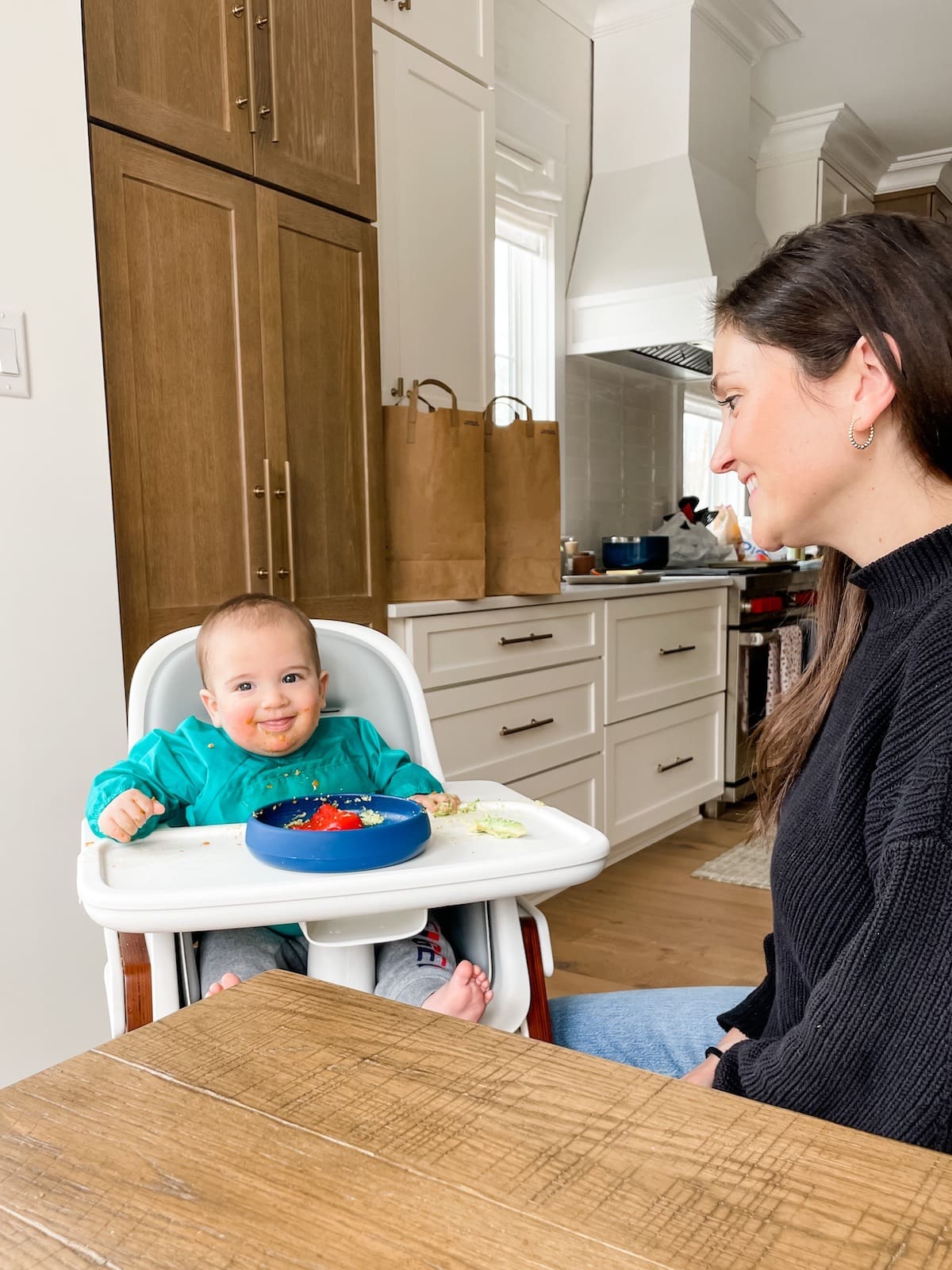 6 month old baby in high chair with blue plate, mom on the side in a black sweater.