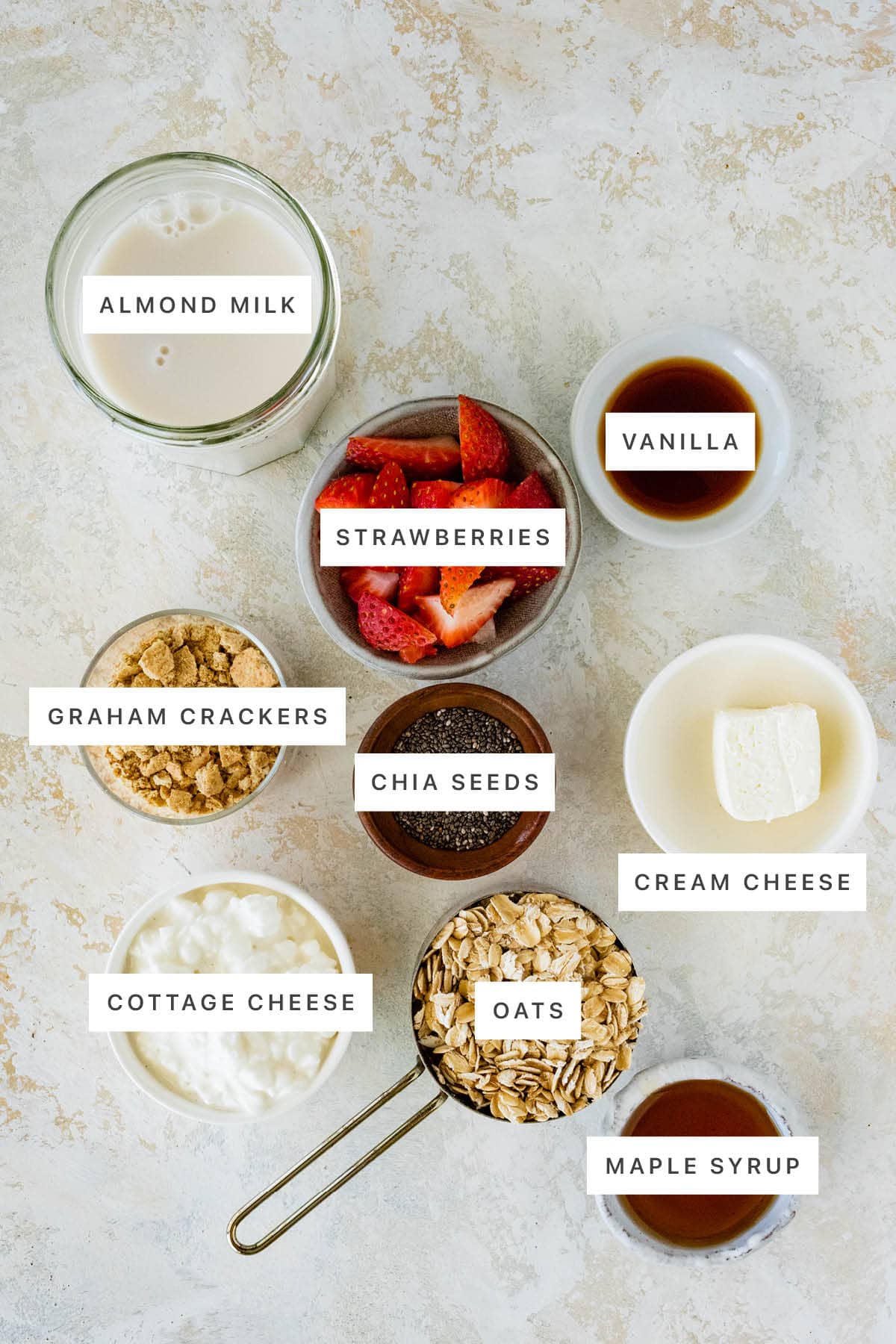 Ingredients measured out to make Strawberry Cheesecake Overnight Oats: almond milk, strawberries, vanilla, graham crackers, chia seeds, cream cheese, cottage cheese, oats and maple syrup.