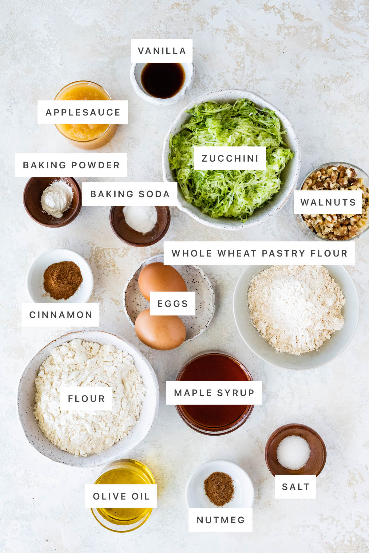 Ingredients measured out to make Healthy Zucchini Bread: vanilla, applesauce, baking powder, baking soda, zucchini, walnuts, cinnamon, eggs, flour, wheat flour, maple syrup, olive oil, nutmeg and salt.