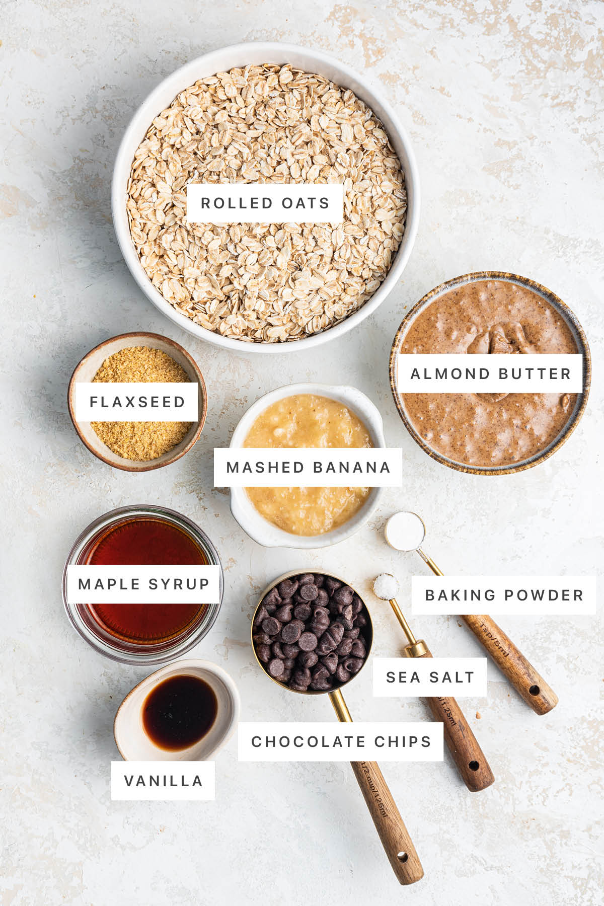 Ingredients measured out to make Healthy Oatmeal Chocolate Chip Bars: rolled oats, almond butter, flaxseed, mashed banana, baking powder, maple syrup, sea salt, chocolate chips and vanilla.