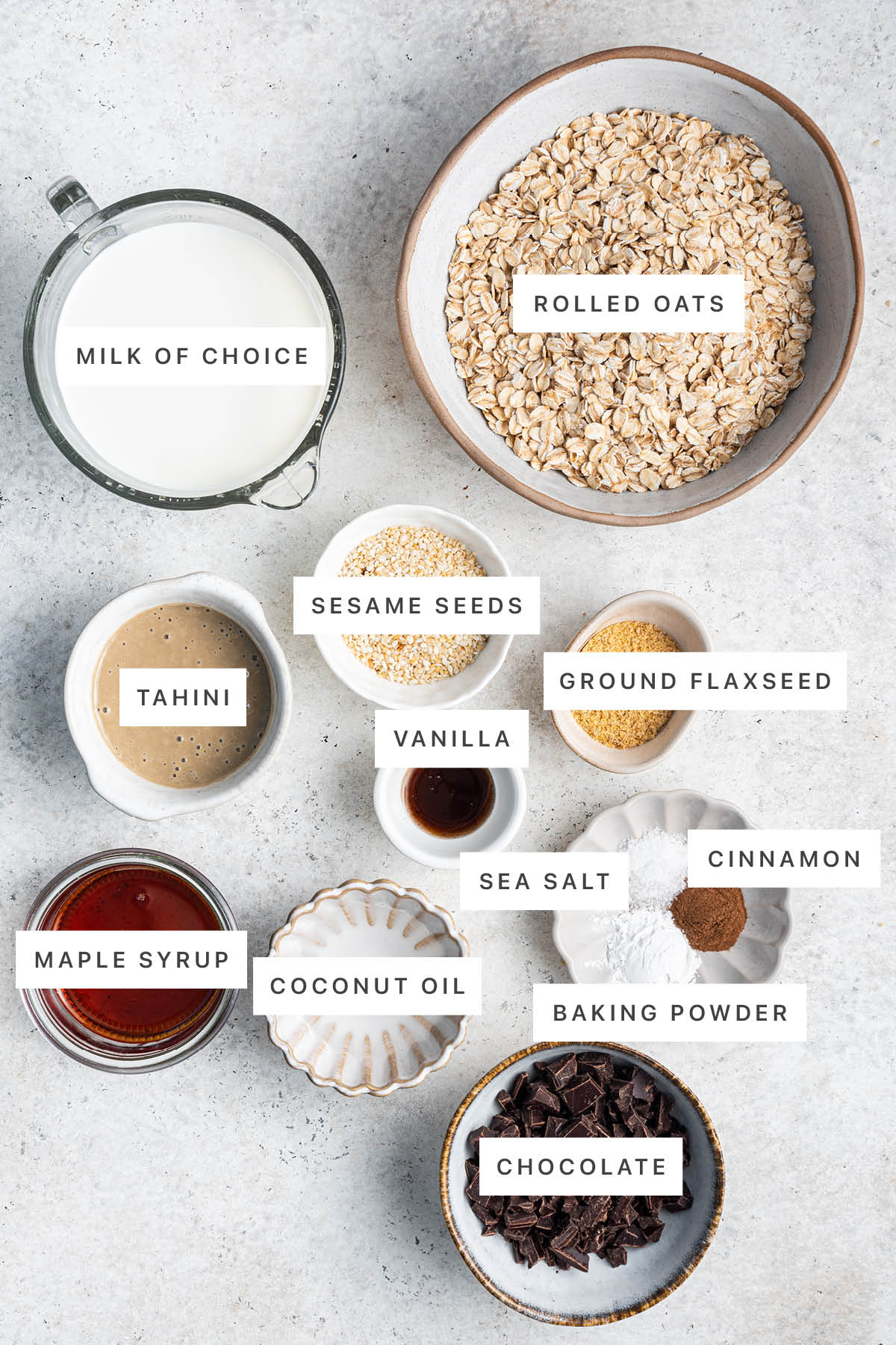Ingredients measured out to make Tahini Baked Oatmeal: milk, rolled oats, tahini, sesame seeds, flaxseed, vanilla, maple syrup, coconut oil, sea salt, cinnamon, baking powder and chocolate.