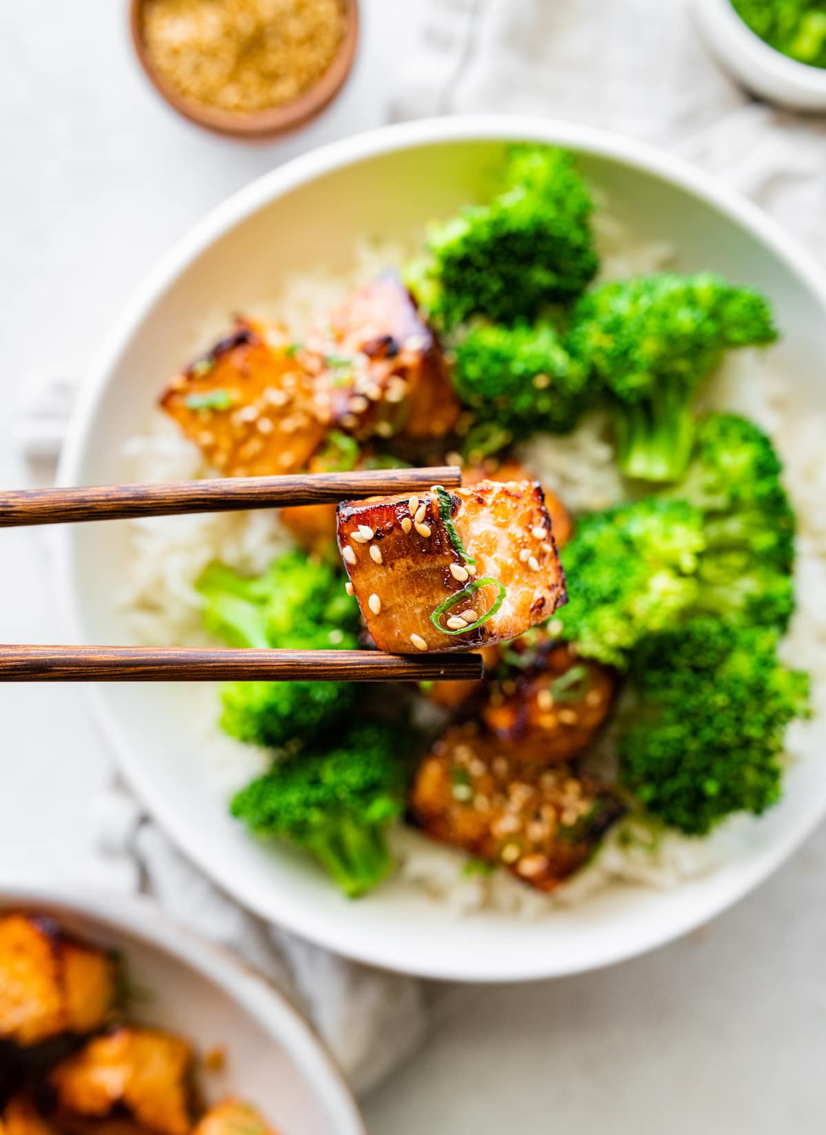 Chopsticks holding one salmon bite over a bowl of salmon bites, broccoli, and rice.