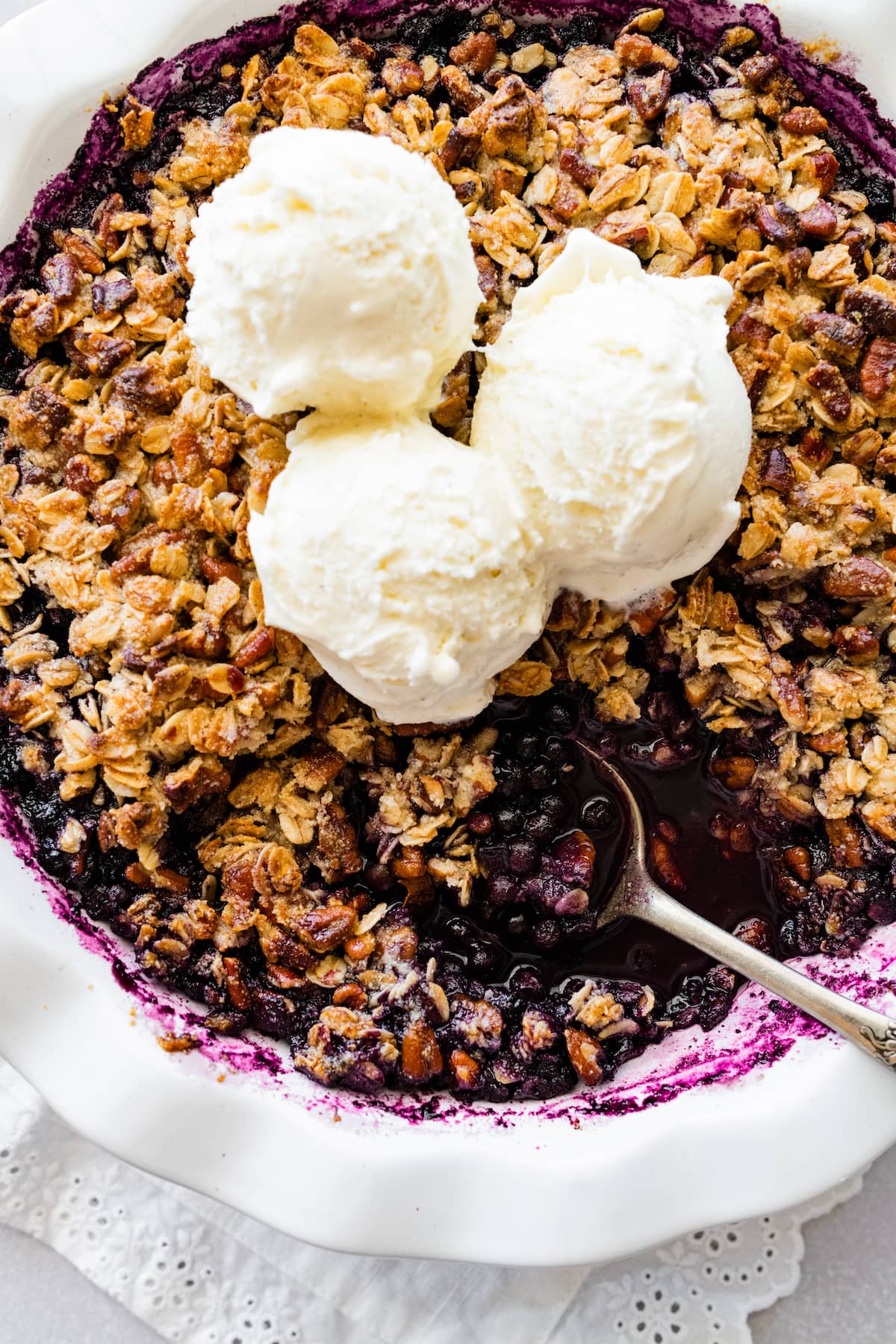 Blueberry Crumble in a pie dish with three scoops of vanilla ice cream on top and a metal spoon.