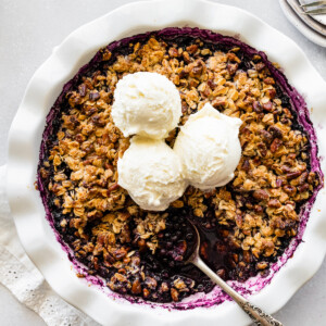 Blueberry crumble in a pie dish with a metal spoon and three scoops of vanilla ice cream.