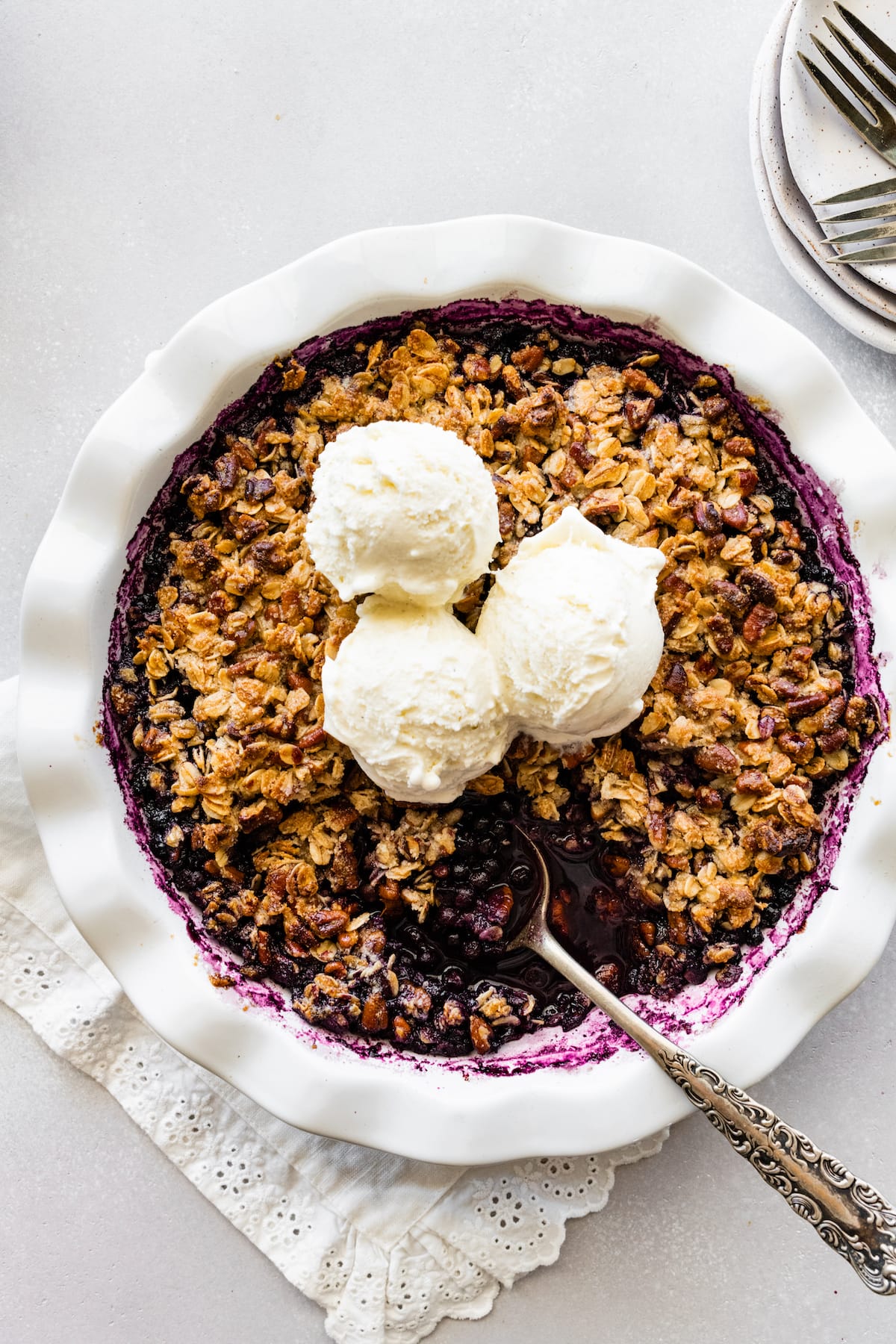 Blueberry crumble in a pie dish with a metal spoon and three scoops of vanilla ice cream.