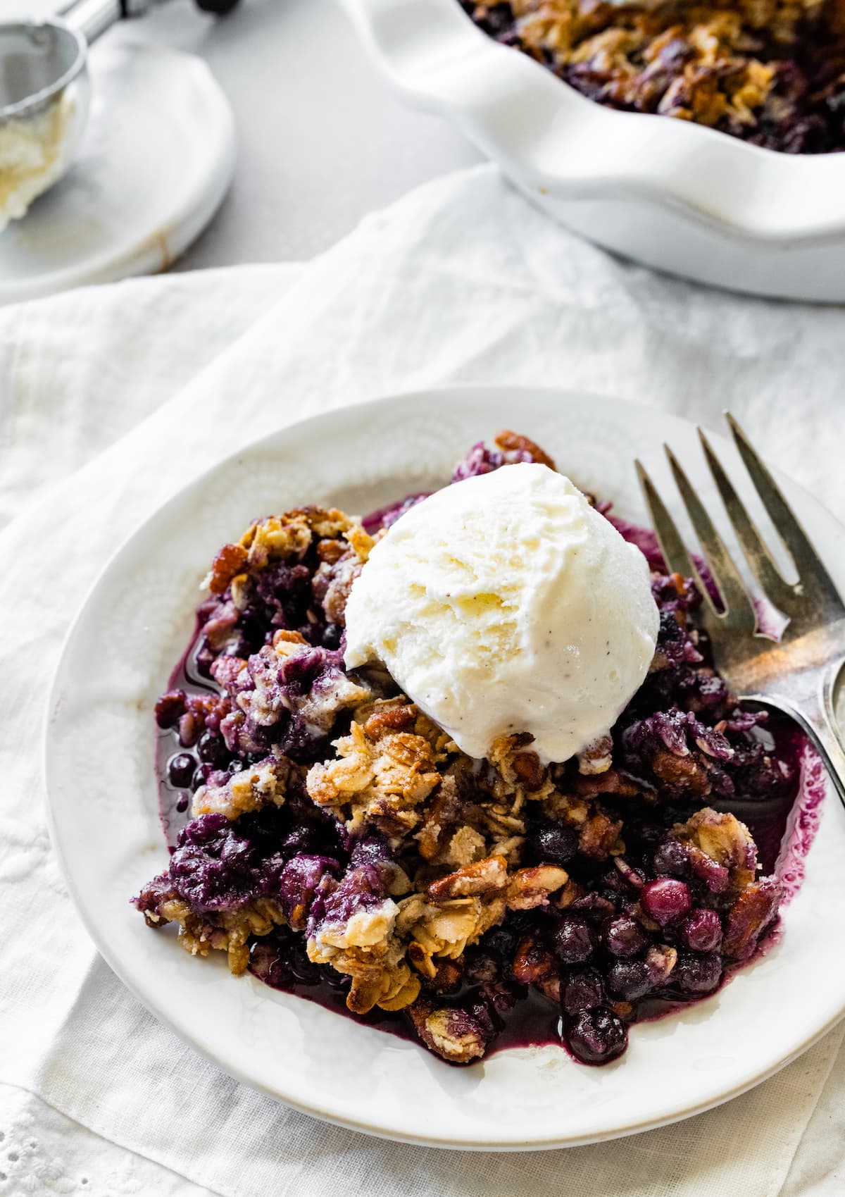 Blueberry crumble in a small bowl with a metal fork and a scoop of vanilla ice cream on top.