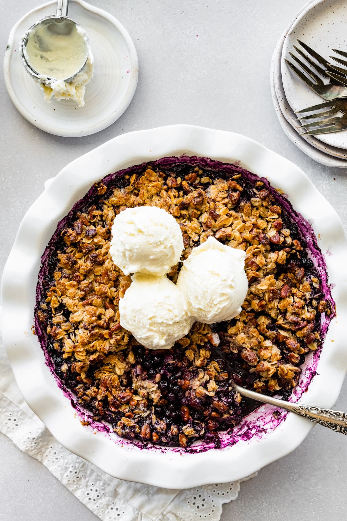 Blueberry Crumble in a pie dish with three scoops of vanilla ice cream on top and a metal spoon.
