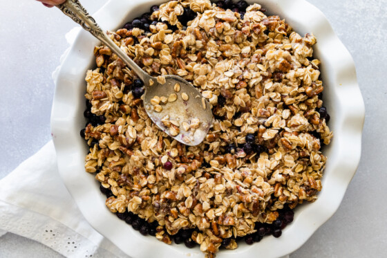 A pie dish with the crumble part being added on top of the blueberries with a metal spoon.