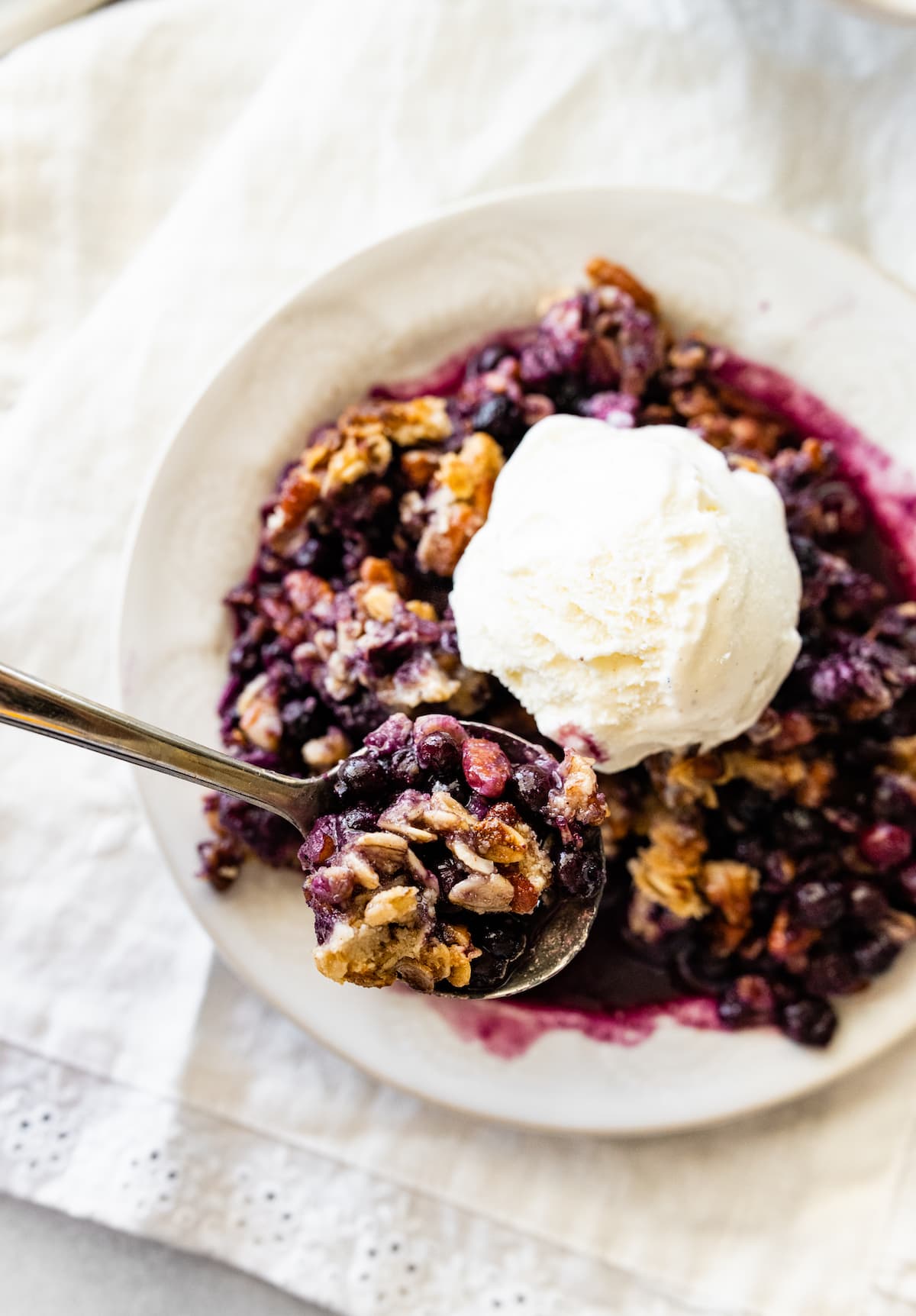 A metal spoon holding a bite size of the blueberry crumble over a small bowl.