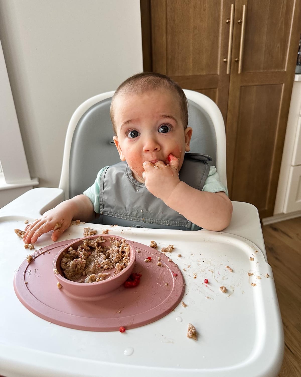 Baby boy eating oatmeal in a high chair with a gray bib and pink bowl.