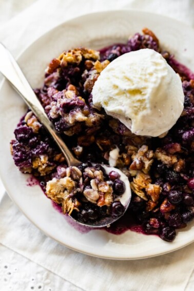 Blueberry crumble in a small bowl with a metal spoon and topped with a scoop of vanilla ice cream.
