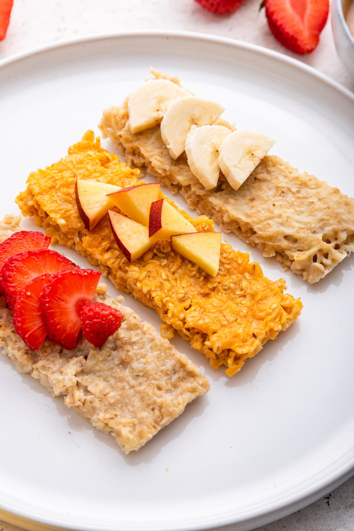 Three oatmeal fingers (banana, sweet potato and applesauce) on a white plate topped with fresh fruit.