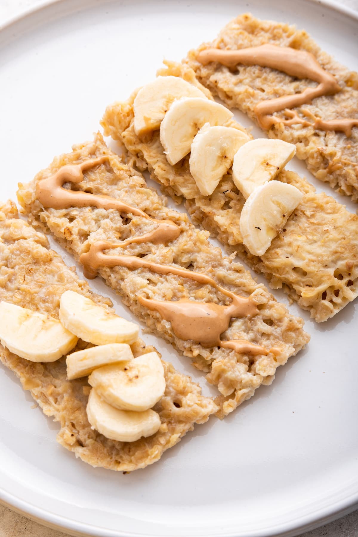 Four banana oatmeal fingers on a white plate topped with almond butter and fresh banana slices.