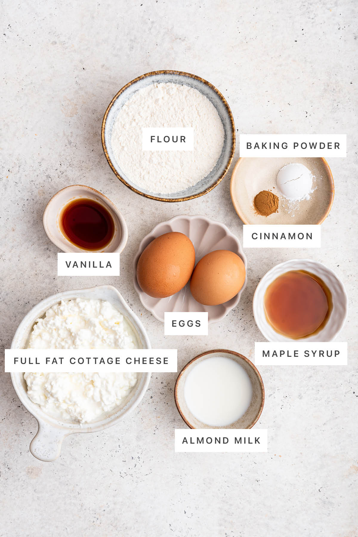 Ingredients measured out to make Cottage Cheese Pancakes: flour, baking powder, cinnamon, vanilla, eggs, maple syrup, cottage cheese and almond milk.