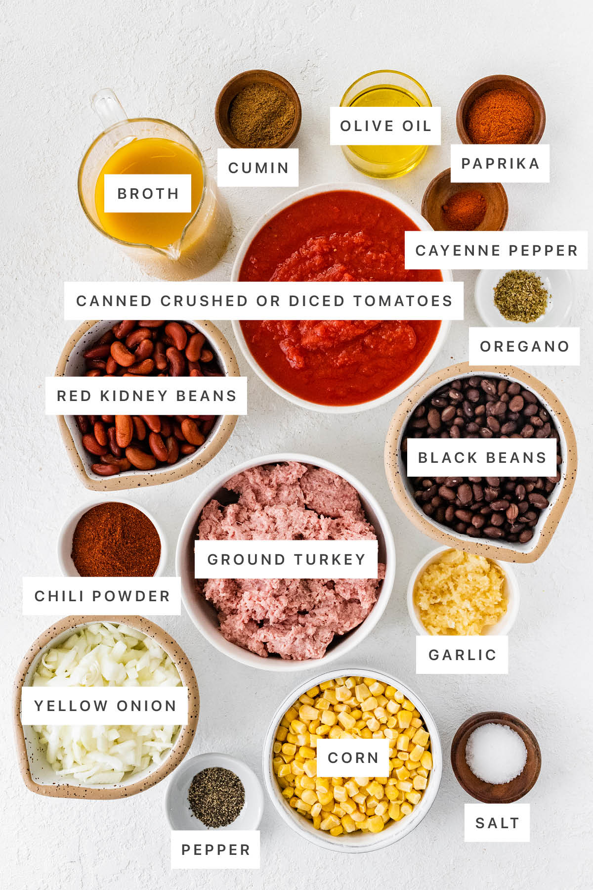 Ingredients measured out to make Turkey Chili: broth, cumin, olive oil, paprika, cayenne pepper, canned crushed tomatoes, oregano, kidney beans, black beans, chili powder, ground turkey, garlic, yellow onion, pepper, corn and salt.
