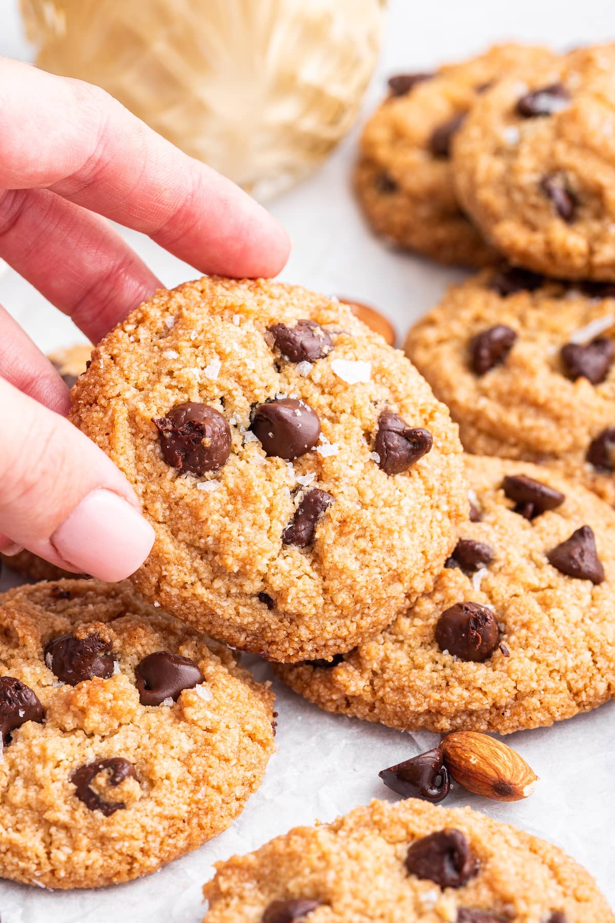 A woman's hand holding an almond flour chocolate chip cookie with multiple other cookies in the background.