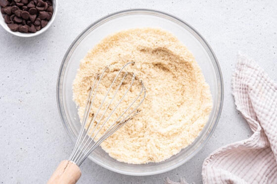 A metal whisk in a large glass bowl with almond flour.