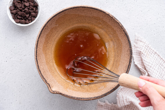 A woman's hand uses a whisk in a mixing bowl of wet ingredients for the almond flour chocolate chip cookies.