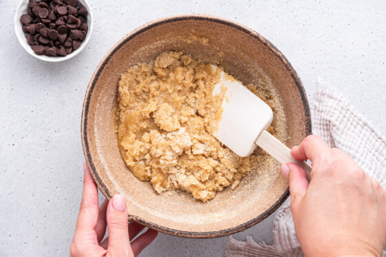 A woman's hand uses a silicone spatula to fold together the batter for almond flour chocolate chip cookies in a large mixing bowl.