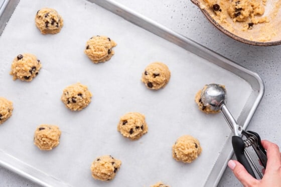 A woman's hand uses a metal ice cream scoop to form the almond flour chocolate chip cookies on a baking tray.