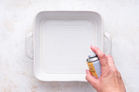 A woman's hand uses a non-stick cooking spray on a square baking dish.