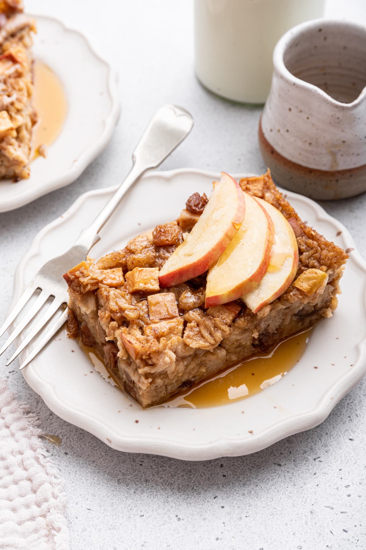 A piece of apple cinnamon baked oatmeal on a small plate topped with sliced apple and maple syrup.