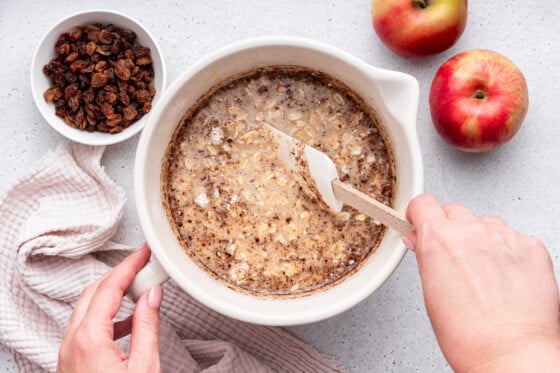 A woman's hand using a silicone spatula to mix the ingredients for the apple cinnamon baked oatmeal in a large mixing bowl.