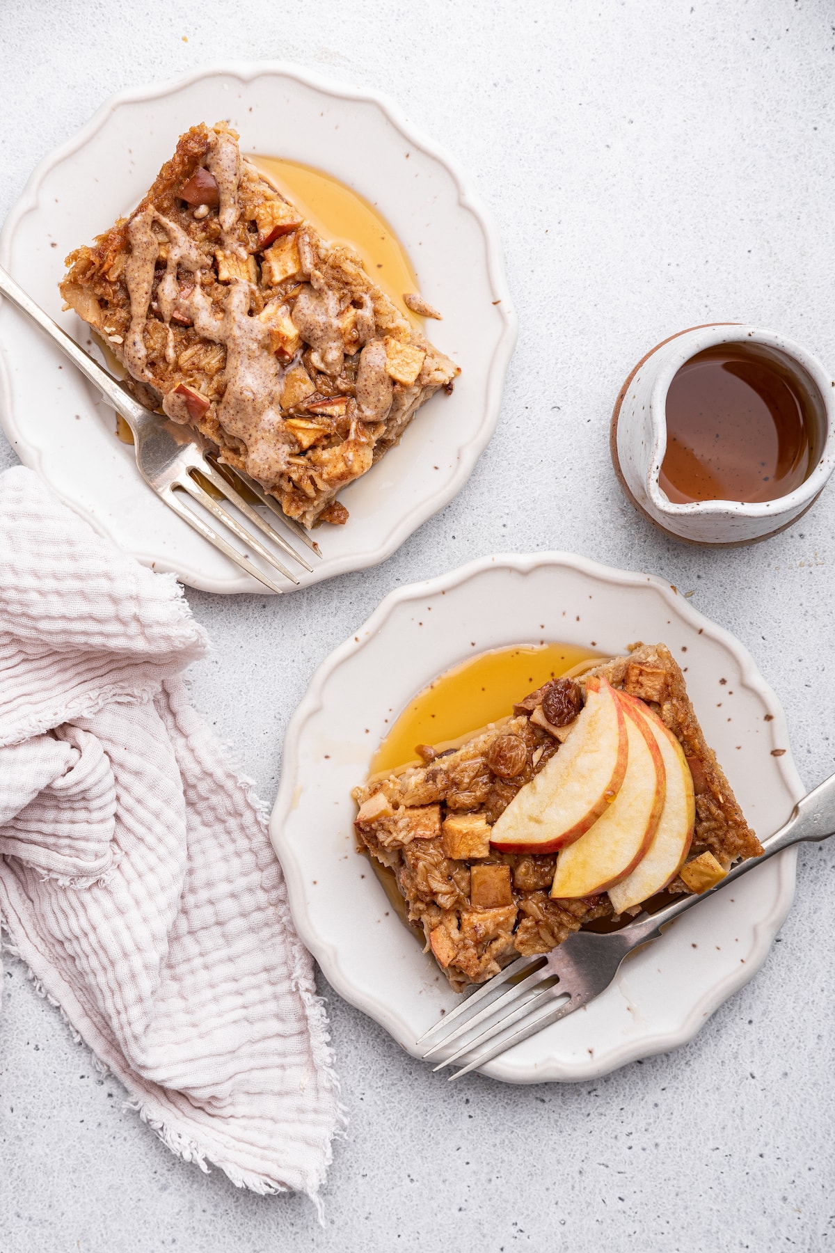 Two slices of apple cinnamon baked oatmeal on separate plates with one slice having sliced apples and maple syrup and the other slice has a drizzle of nut butter.
