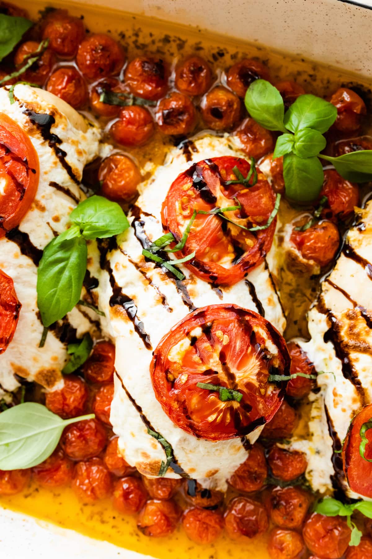 Close up of chicken breasts covered in mozzarella cheese, tomato slices, and balsamic glaze in a rectangular baking dish with cherry tomatoes and fresh basil.