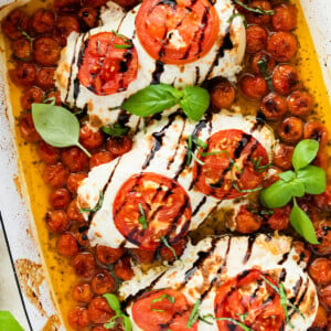 Three chicken breasts covered in mozzarella cheese, tomato slices, and balsamic glaze in a rectangular baking dish with cherry tomatoes and fresh basil.